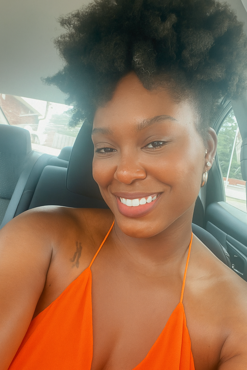 Woman in an orange top sitting inside a car, smiling.